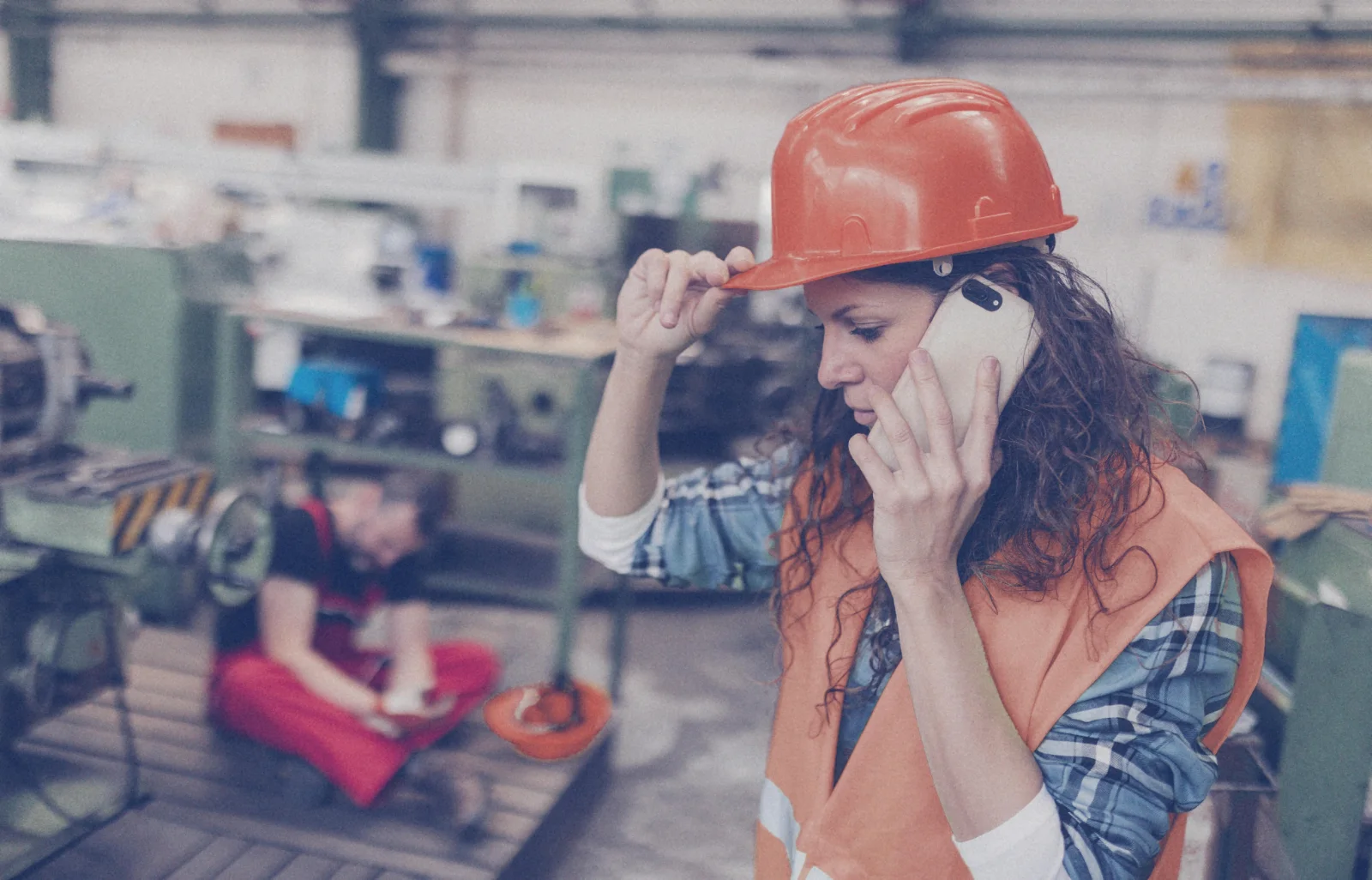 A worker in a hard hat and safety vest takes a phone call on the factory floor, with a colleague in the background looking at a tablet.